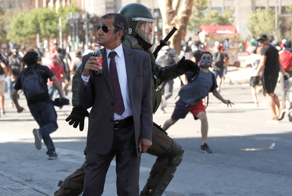 A man drinks a Frucola as a policeman chases demonstrators during a protest against Chile's government, in Santiago, Chile, on December 4, 2019.jpg